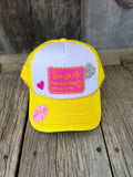 A yellow and white trucker hat with a pink embroidered patch and decorative pink and silver heart embellishments, displayed on a rustic wooden surface.