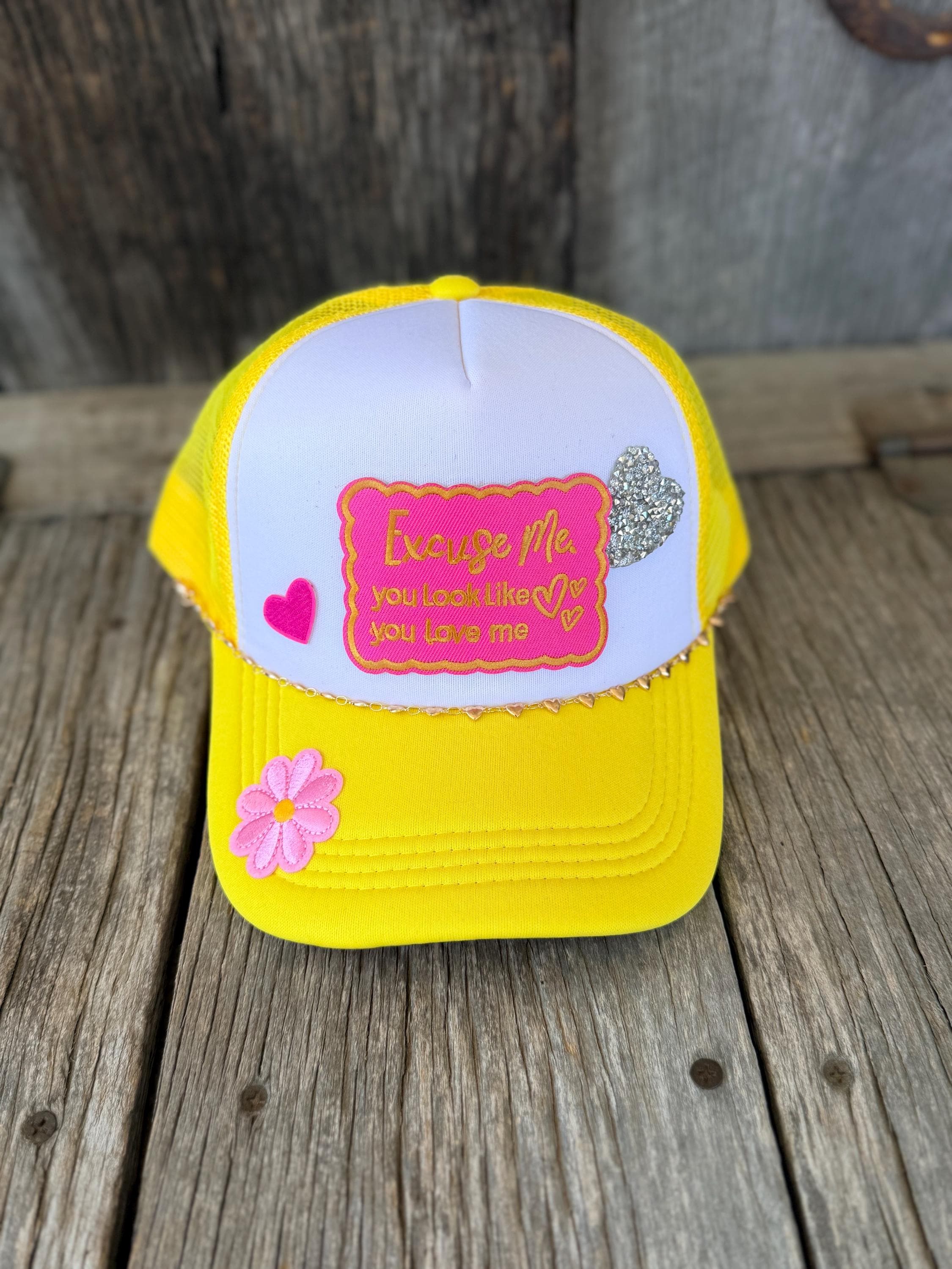 A yellow and white trucker hat with a pink embroidered patch and decorative pink and silver heart embellishments, displayed on a rustic wooden surface.