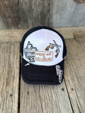 A black and white trucker hat with embroidered graphics on a rustic wooden surface. The front panel is white with black mesh, featuring a brown and white 'Whiskey patch and cowboy/horse embroidery.