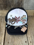 A black and white trucker hat with embroidered 'Howdy' and 'Cowboy' text, a cowboy hat patch, and dangling silver charms, displayed on a rustic wooden surface.