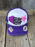 A purple and white trucker hat with mesh back, decorated with a silver rhinestone chain, a black patcha pink glitter star, a rhinestone heart patch, and a 'DARLIN' patch, displayed on a rustic wooden surface.