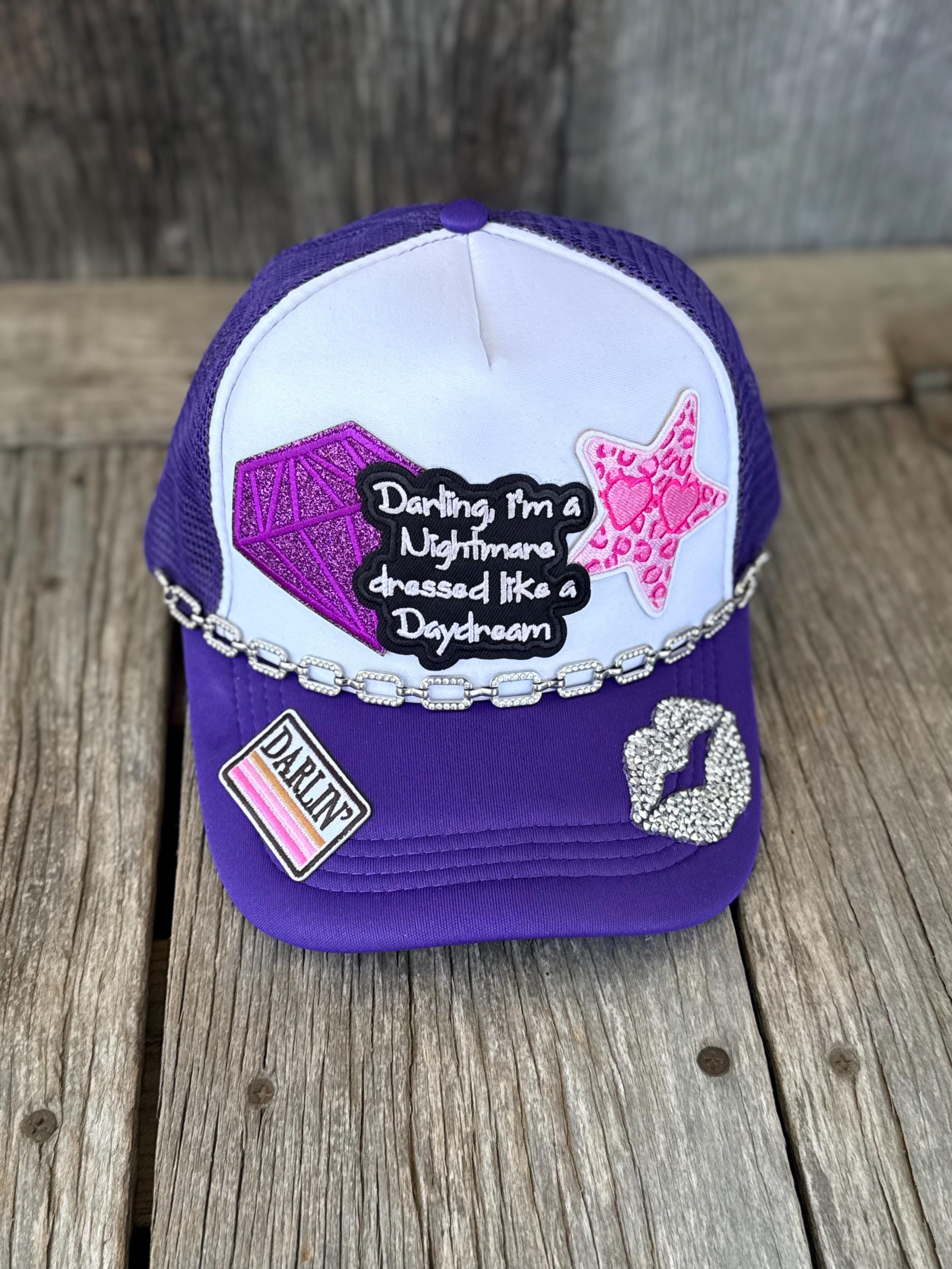A purple and white trucker hat with mesh back, decorated with a silver rhinestone chain, a black patcha pink glitter star, a rhinestone heart patch, and a 'DARLIN' patch, displayed on a rustic wooden surface.