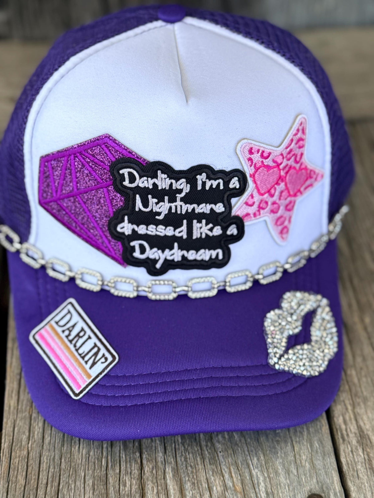 A purple and white trucker hat with custom rhinestone patches, including a black text patch, a pink leopard print star, a pink diamond, and a rhinestone lip print, displayed on a rustic wooden surface.