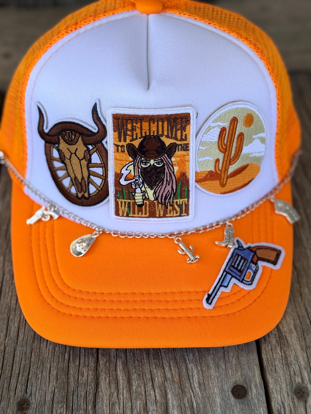 An orange and white trucker hat with three embroidered patches on the front panel and silver dangling charms, displayed on a rustic wooden surface.