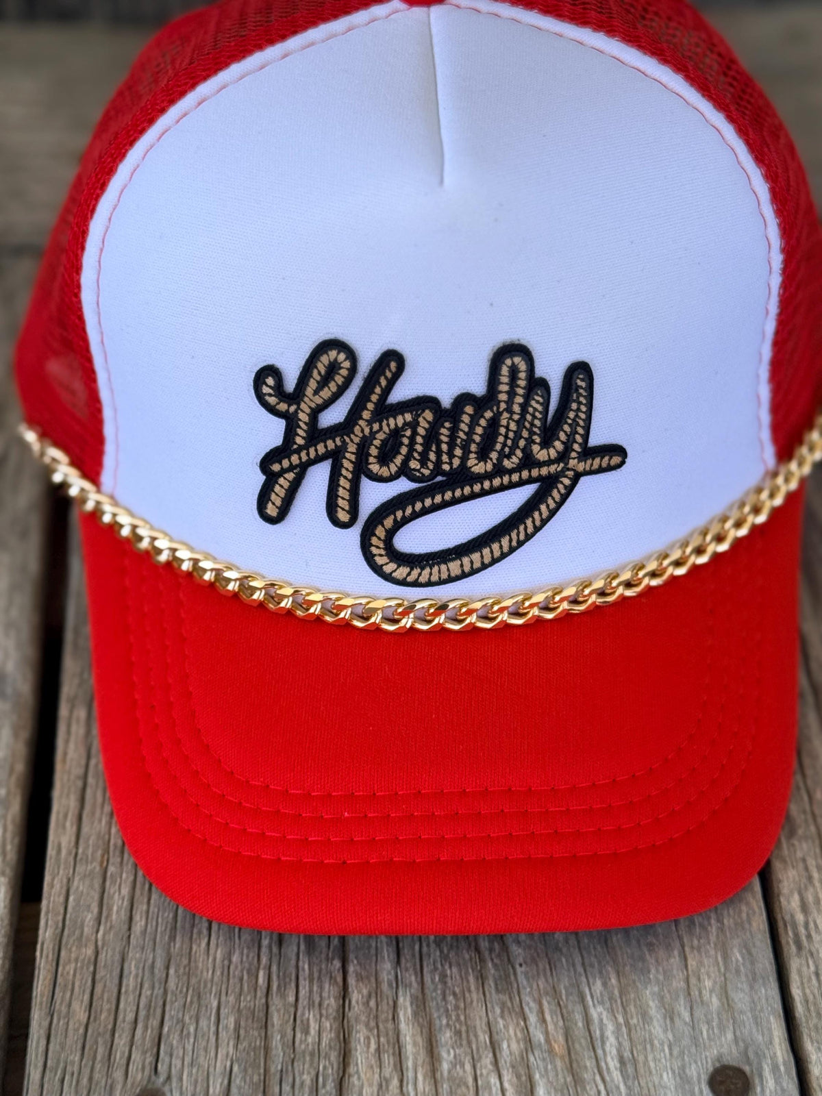 A red and white trucker hat with a gold chain across the front and black embroidered script reading 'Hardy' on a white front panel, photographed on a rustic wooden surface.