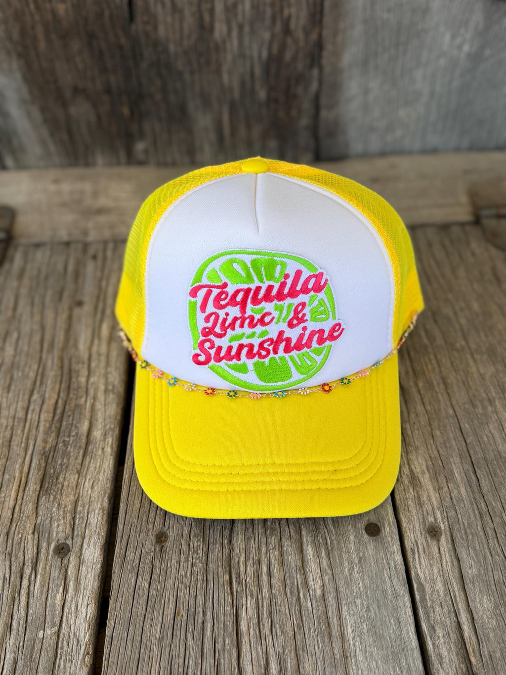 A yellow and white trucker hat with a graphic on the front that reads 'Tequila Lime & Sunshine' in pink and green, resting on a rustic wooden surface.