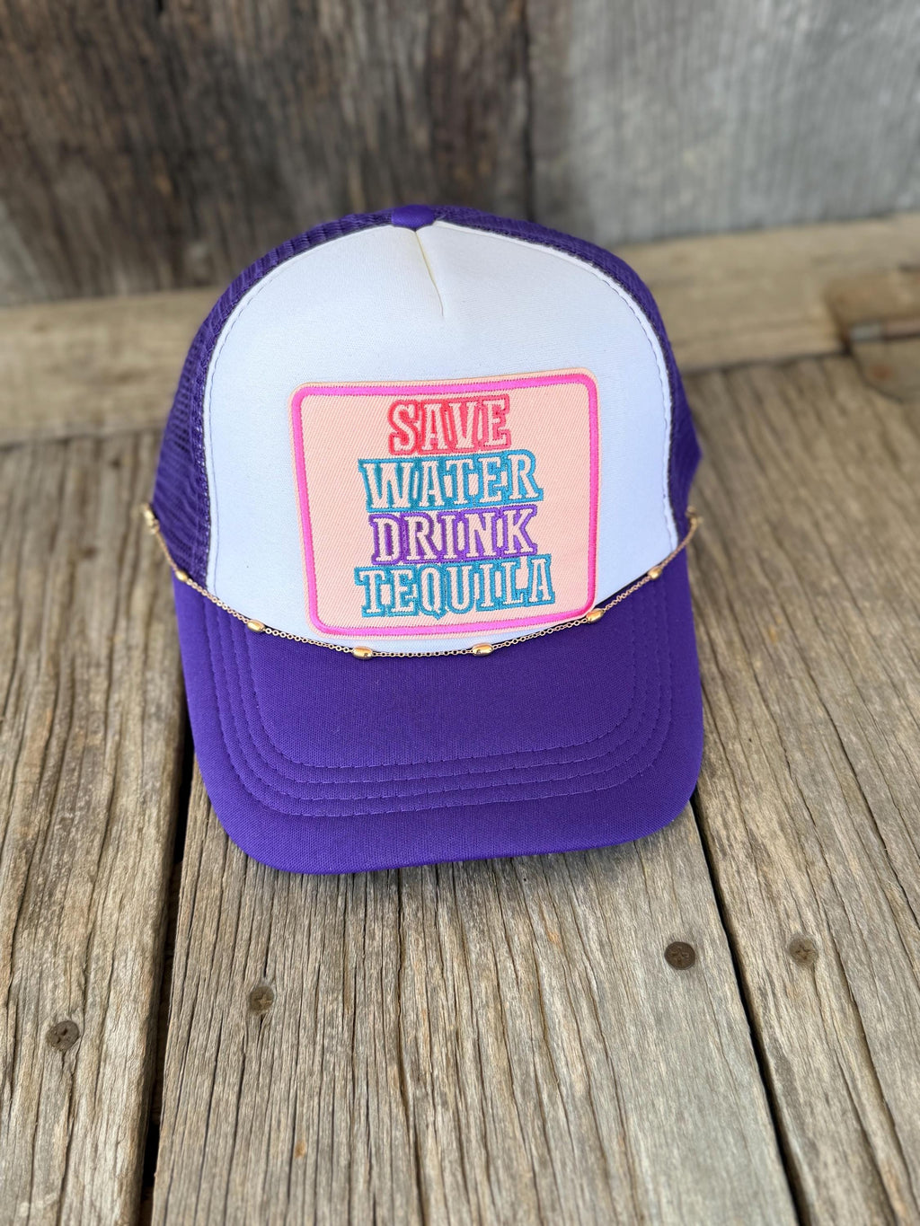 A purple and white trucker hat with a pink and blue embroidered patch reading 'SAVE WATER DRINK TEQUILA!' resting on a rustic wooden surface.