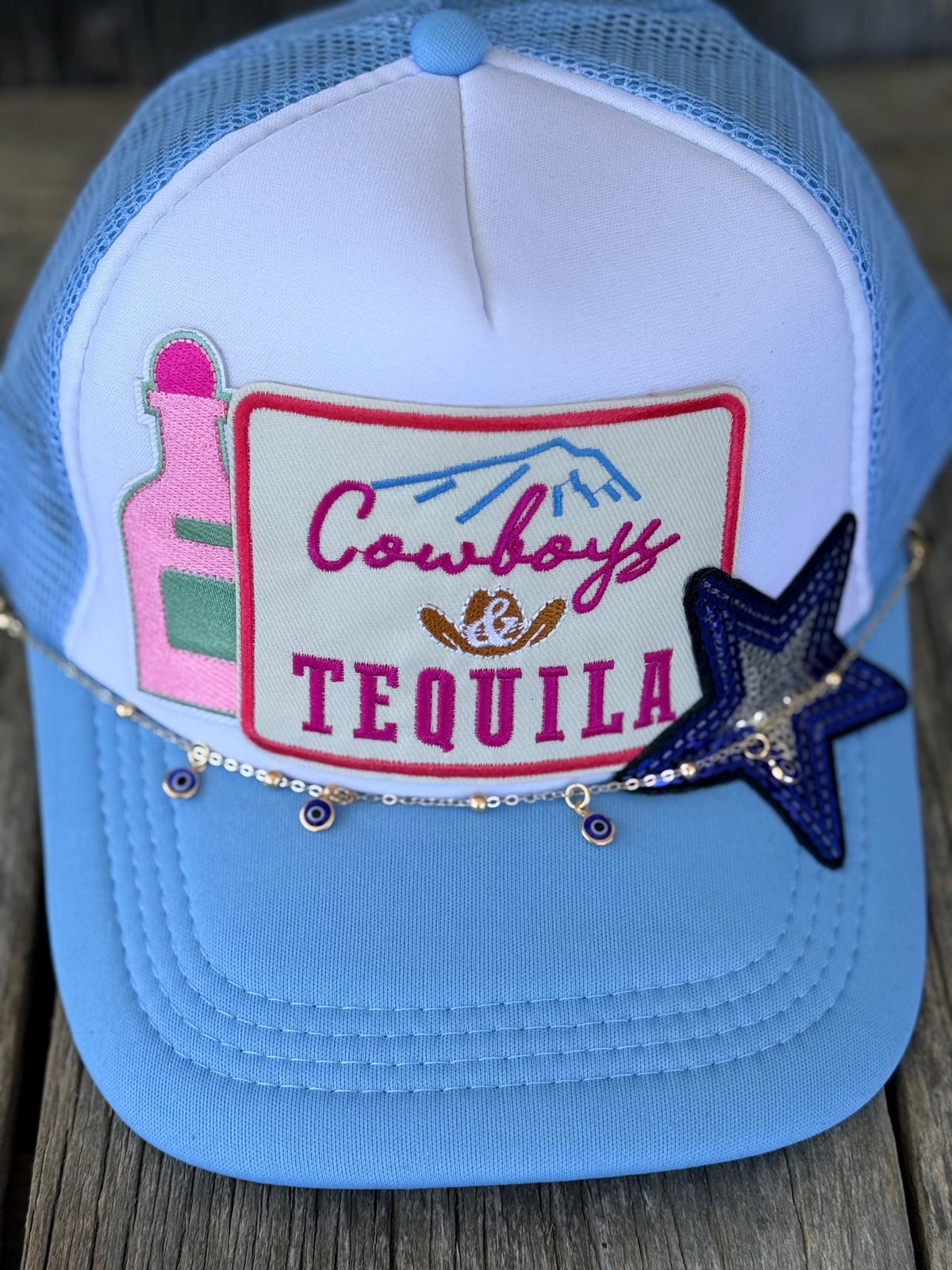 A light blue and white trucker hat with a front patch reading 'Cowboys & Tequila' and decorative embroidered details, photographed on a rustic wooden surface.