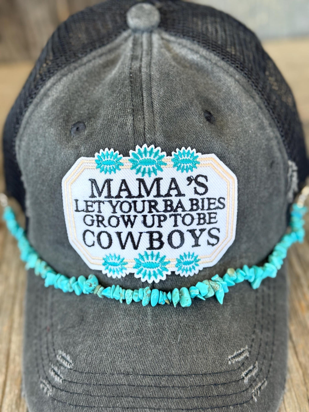 A distressed dark gray trucker hat with a black mesh back, featuring a white embroidered patch that reads 'MAMA'S LET YOUR BABIES GROW UP TO BE COWBOYS' surrounded by turquoise floral designs and a matching turquoise beaded hat chain.