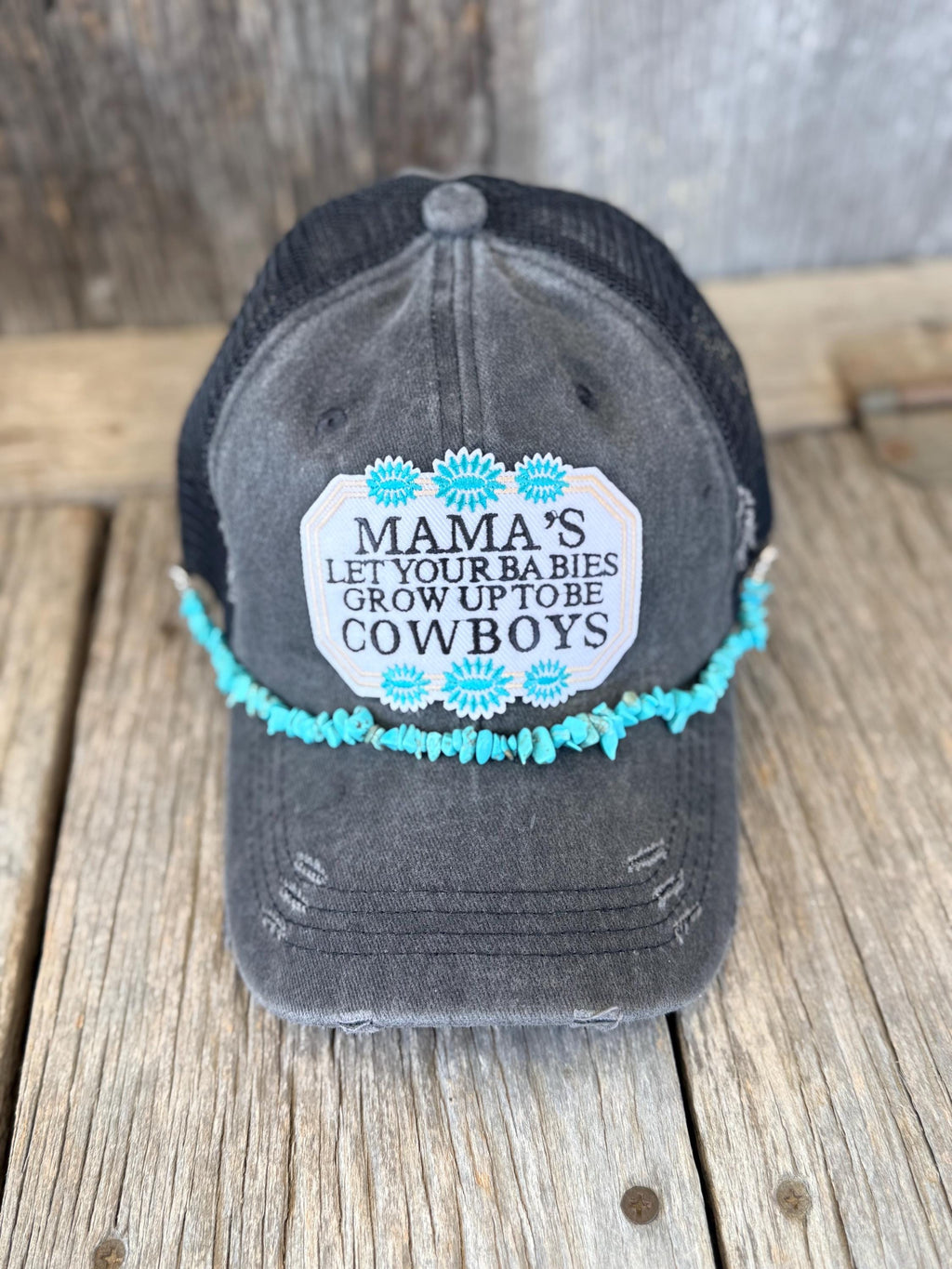 A distressed dark gray trucker hat with a black mesh back, featuring a white embroidered patch' surrounded by turquoise flowers, and a turquoise beaded hat chain, displayed on a rustic wooden surface.