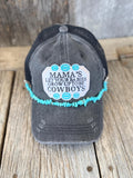 A distressed dark gray trucker hat with a black mesh back, featuring a white embroidered patch' surrounded by turquoise flowers, and a turquoise beaded hat chain, displayed on a rustic wooden surface.