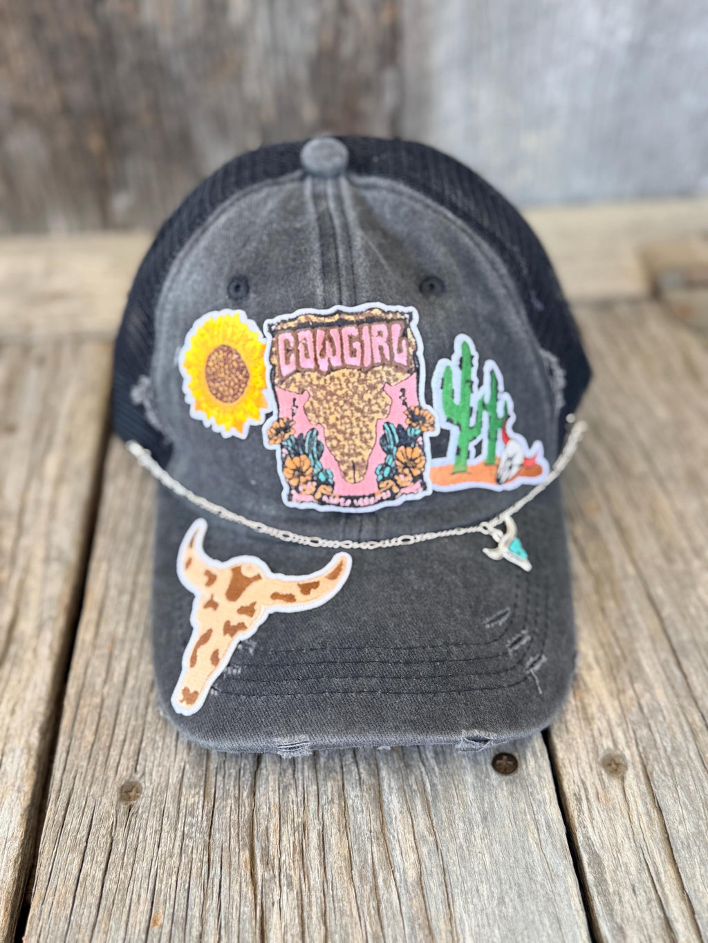 A distressed black trucker cap with a weathered gray finish, decorated with a variety of colorful embroidered patches including a sunflower, a pink 'Cowgirl' patch, a cactus, and a longhorn steer patch. The cap is displayed on a rustic wooden surface.