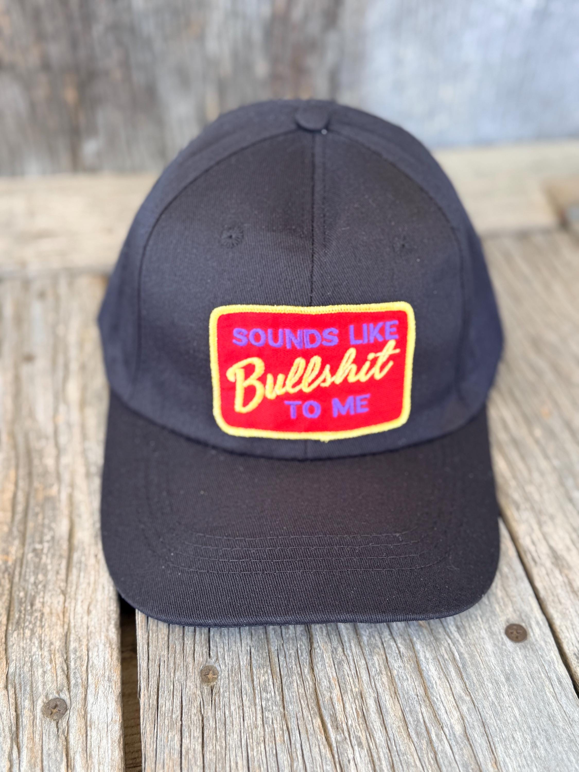 A black baseball cap with a rectangular embroidered patch on the front that reads 'SOUNDS LIKE Bullshit TO ME' in red and yellow, displayed on a rustic wooden surface.