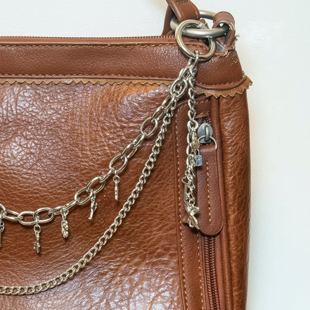 Close-up detail of gold-tone oval link chain and delicate curb chain clipped to tan distressed Bandana leather crossbody bag hardware with key and padlock charms hanging from side zipper pull, Western boho handbag jewelry