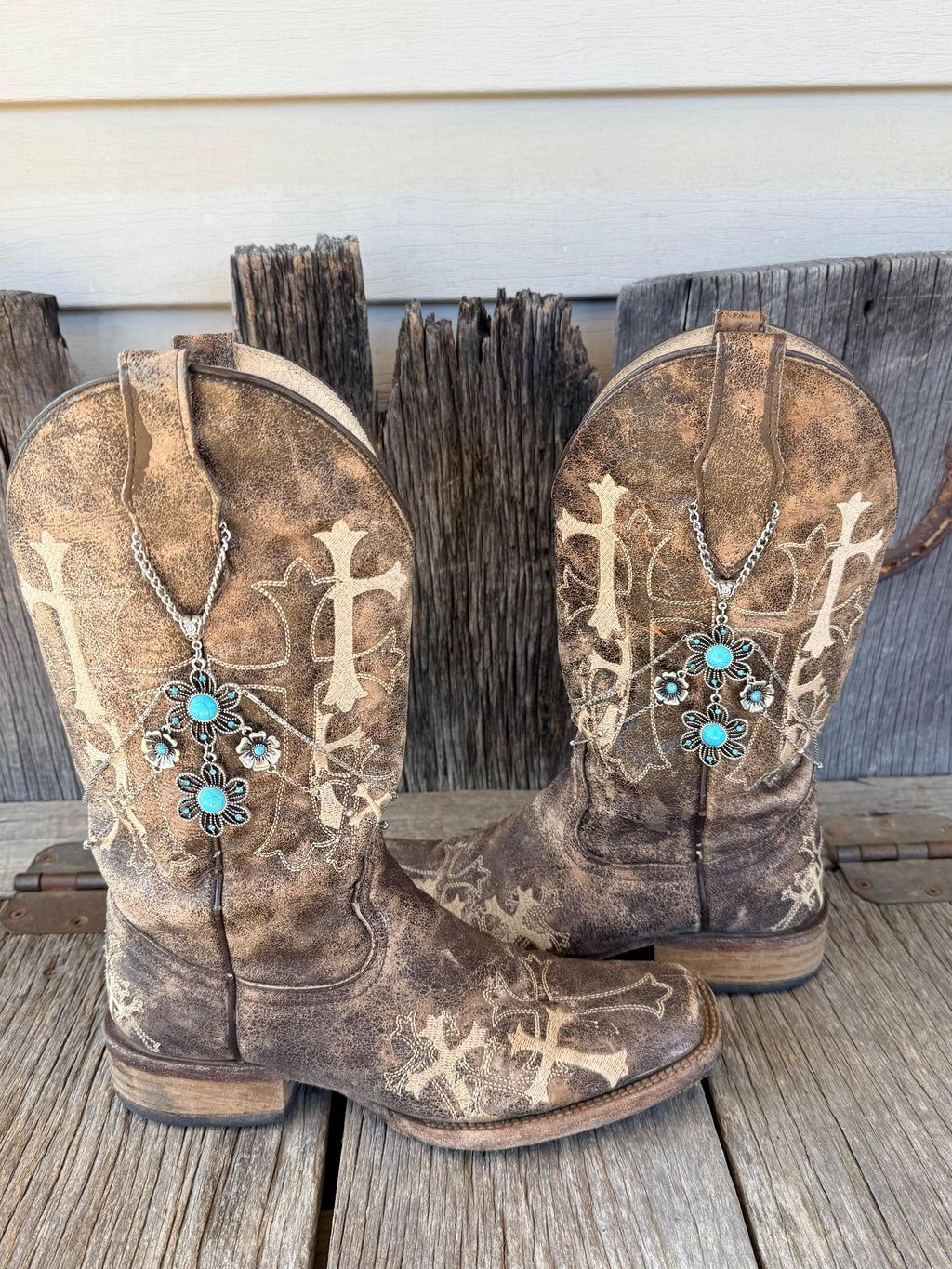 Angled side view of pair of distressed brown cross embroidered cowboy boots against weathered wood fence with silver multi-chain boot bracelets and turquoise stone flower charm clusters