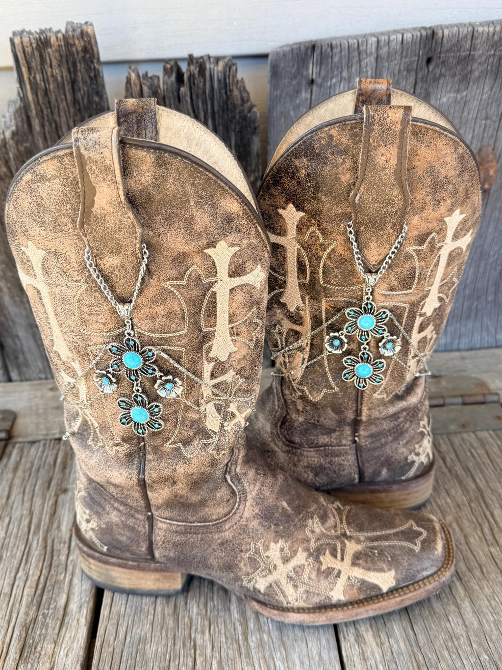 Front view of pair of distressed brown cross embroidered cowboy boots on rustic wood planks wearing silver multi-chain boot bracelets with turquoise stone flower charm clusters, set of 2