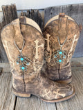 Front view of pair of distressed brown cross embroidered cowboy boots on rustic wood planks wearing silver multi-chain boot bracelets with turquoise stone flower charm clusters, set of 2