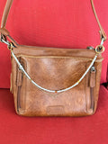 Front view of tan distressed American West leather crossbody bag against red background with two-strand Western boho purse necklace featuring white seed bead strand with red heart bead accent and gold heart bead accents above, and delicate gold cable chain with small open gold heart pendant below, clipped via gold rings