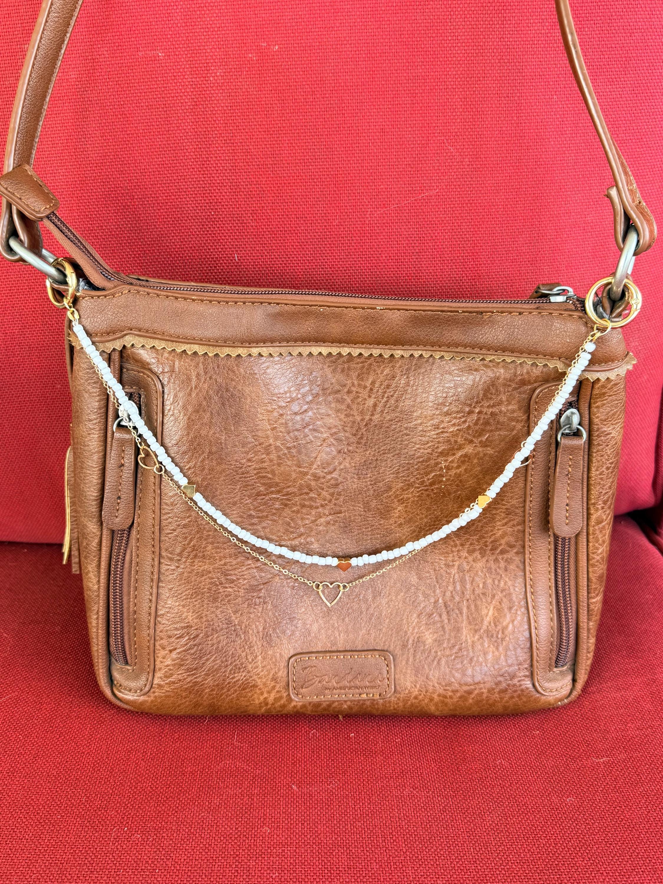 Front view of tan distressed American West leather crossbody bag against red background with two-strand Western boho purse necklace featuring white seed bead strand with red heart bead accent and gold heart bead accents above, and delicate gold cable chain with small open gold heart pendant below, clipped via gold rings