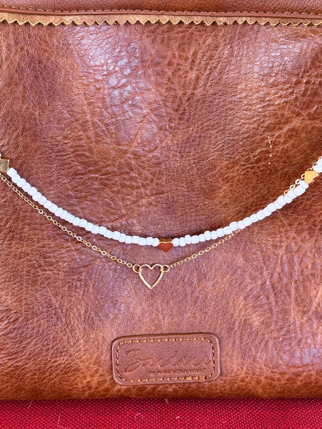 Close-up of tan distressed American West leather crossbody bag showing white seed bead strand with single red heart bead accent at center and gold heart bead accents near ends above, and delicate gold cable chain with small open gold heart pendant centered below, Western boho purse necklace detail