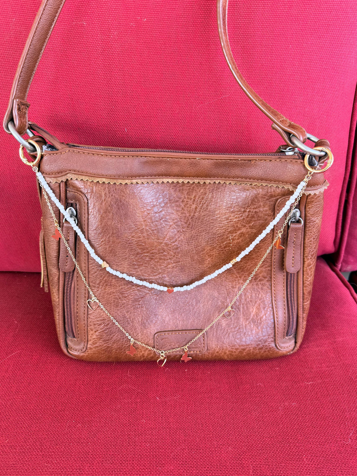 Front view of tan distressed American West leather crossbody bag against red background with two-strand Western boho purse necklace featuring white seed bead strand with red heart bead accent and gold tube bead accents above, and delicate gold cable chain with two gold butterfly charms and open gold heart pendant below