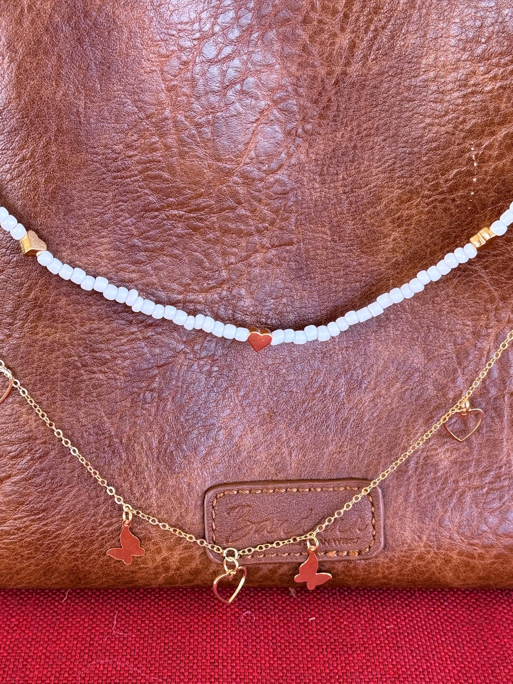 Close-up of tan distressed American West leather crossbody bag showing white seed bead strand with single red heart bead accent at center and gold rectangular tube bead accents near ends above, and delicate gold cable chain with two gold butterfly charms and open gold heart pendant centered below, Western boho purse necklace detail