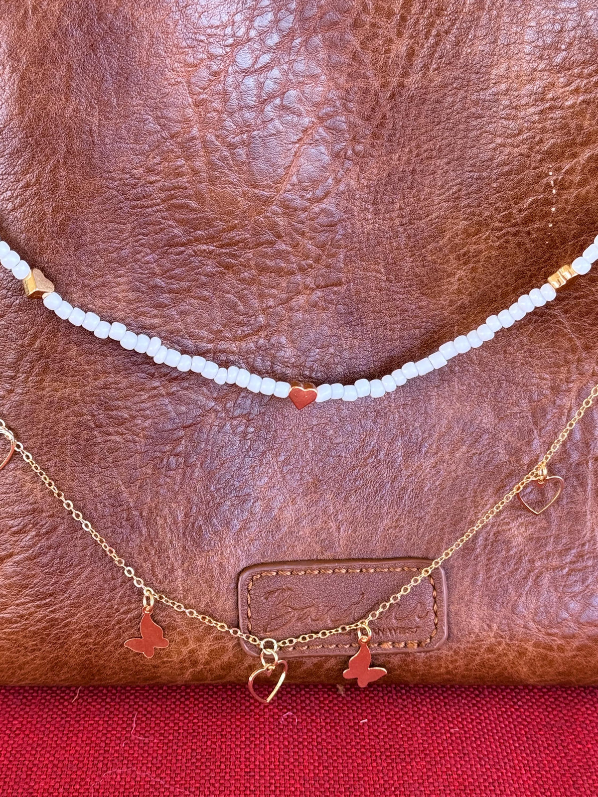 Close-up of tan distressed American West leather crossbody bag showing white seed bead strand with single red heart bead accent at center and gold rectangular tube bead accents near ends above, and delicate gold cable chain with two gold butterfly charms and open gold heart pendant centered below, Western boho purse necklace detail