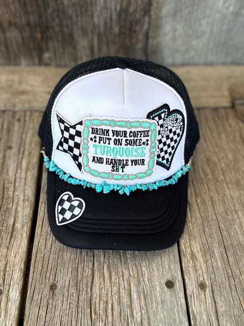 A black and white trucker hat with turquoise fringe and a front patch that reads 'DRINK YOUR COFFEE PUT ON SOME TURQUOISE AND HANDLE YOUR SH*T'. The hat is photographed on a rustic wooden surface.
