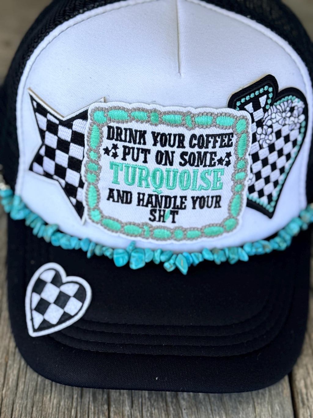 A trucker hat with a white front panel and black brim, decorated with a turquoise and black checkered patch that reads 'DRINK YOUR COFFEE PUT ON SOME TURQUOISE AND HANDLE YOUR SH*T'. The brim also features a small black-and-white checkered heart patch and a turquoise beaded trim.