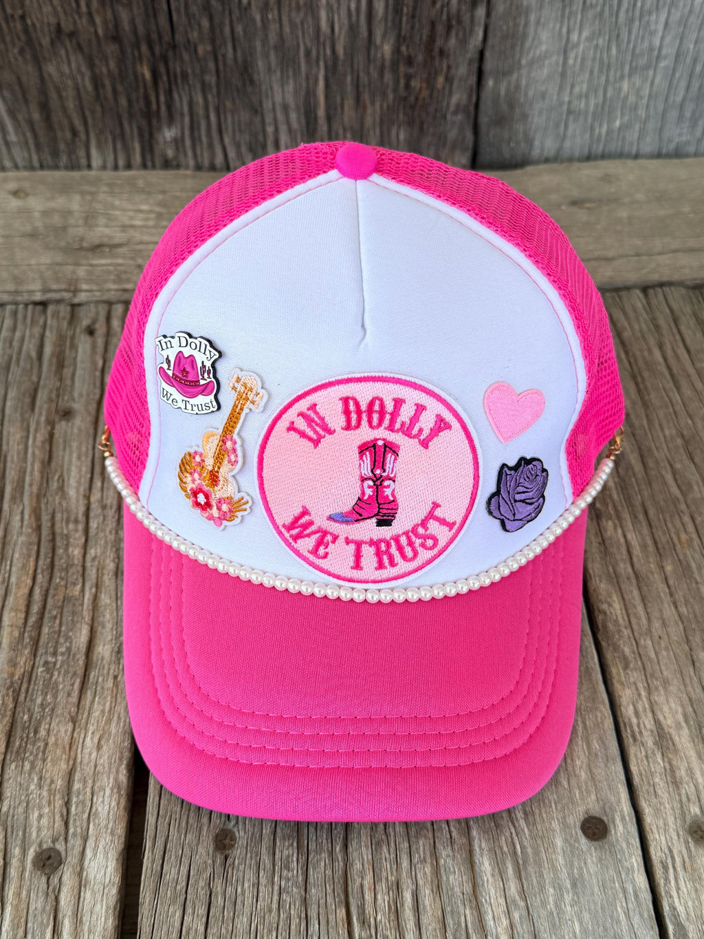 A pink and white trucker hat with mesh back panels, decorated with embroidered patches including a guitar, a pink heart, a purple rose, and a central circular patch'. The hat is displayed on a rustic wooden plank surface.