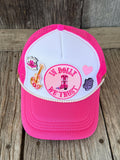 A pink and white trucker hat with mesh back panels, decorated with embroidered patches including a guitar, a pink heart, a purple rose, and a central circular patch'. The hat is displayed on a rustic wooden plank surface.