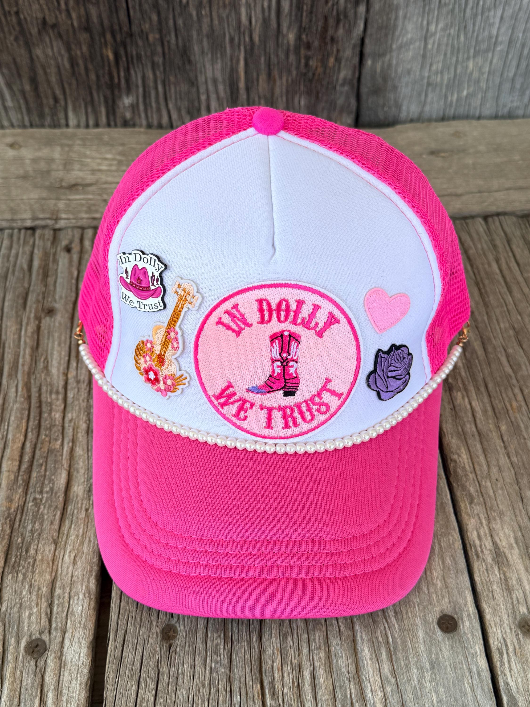 A pink and white trucker hat with mesh back panels, decorated with embroidered patches including a guitar, a pink heart, a purple rose, and a central circular patch'. The hat is displayed on a rustic wooden plank surface.