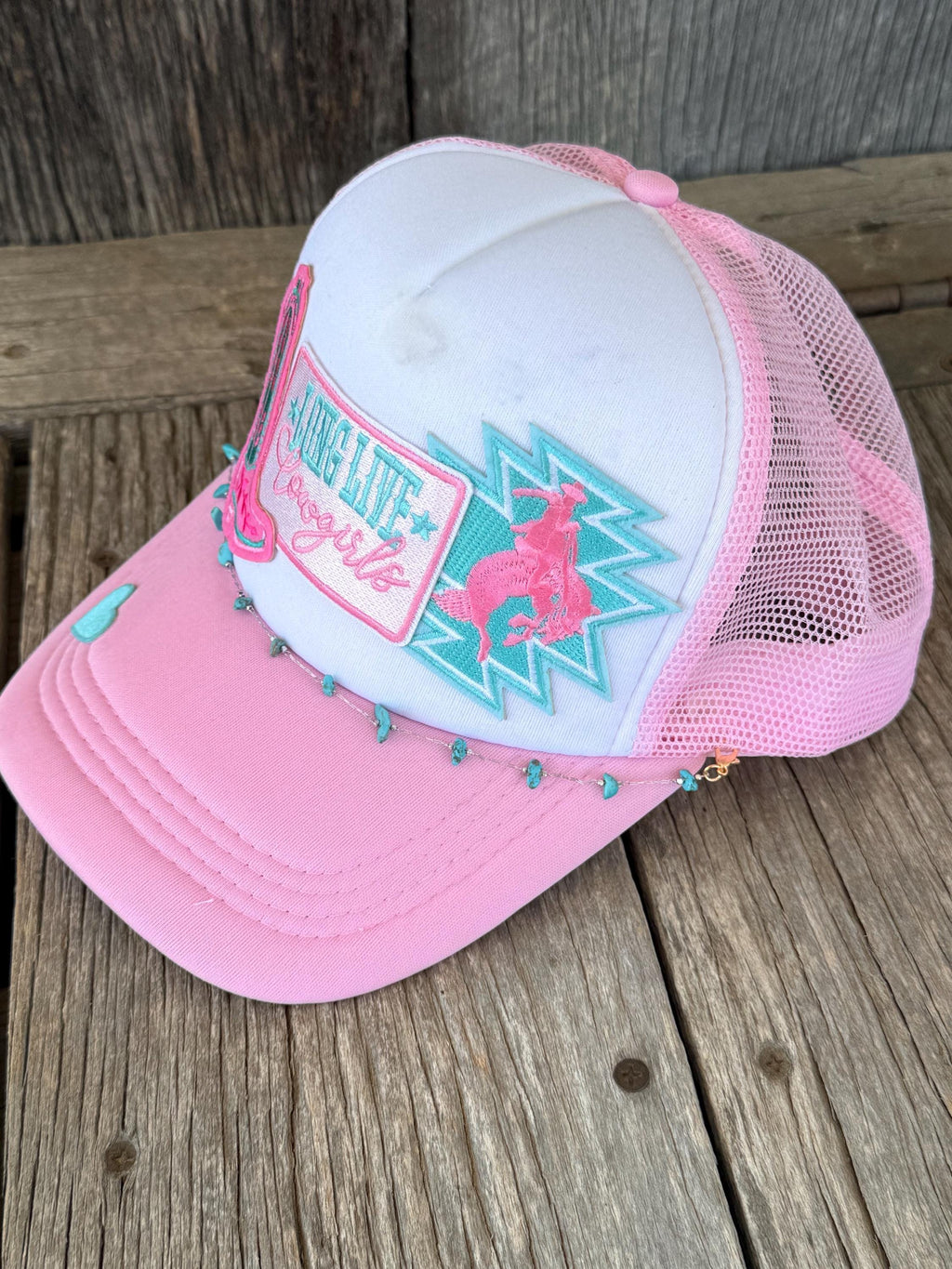 A pink and white trucker hat with embroidered patches, resting on a rustic wooden surface. The hat features a white front panel, a pink brim, and pink mesh back panels.