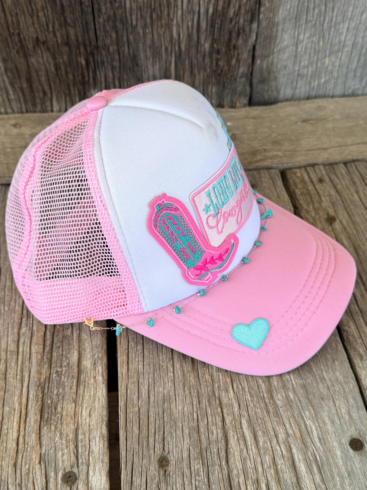 Pink and white trucker hat with mesh back, featuring a pink cowboy hat patch with turquoise details, a pink and white patch, turquoise rhinestone accents, and a light blue heart on the brim, displayed on a rustic wooden surface.