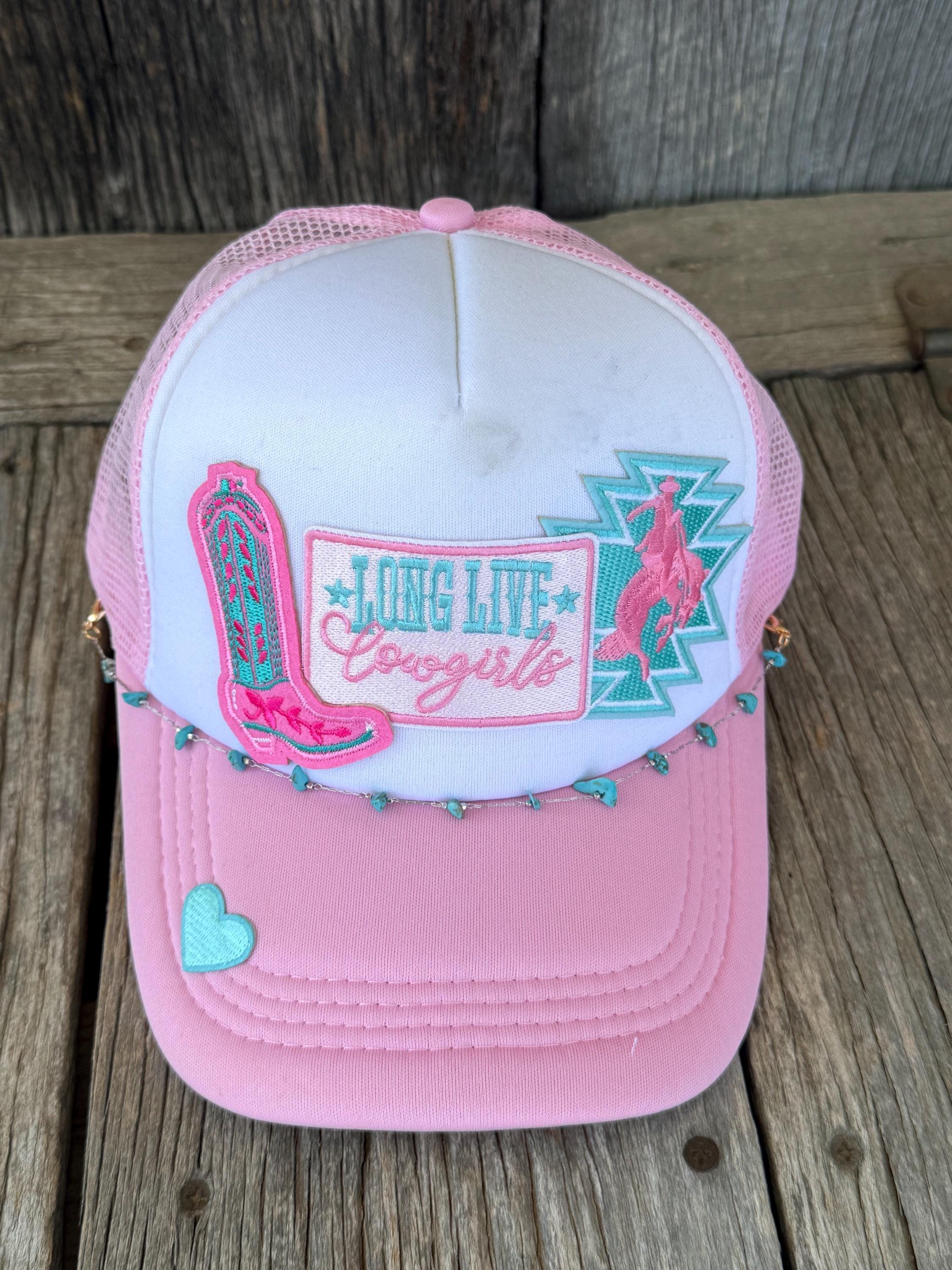 A pink and white trucker hat with western-themed embroidered patches, including a cowboy boot, a cowboy silhouette, and the text 'LONG Live Cowgirls,' displayed on a rustic wooden surface.