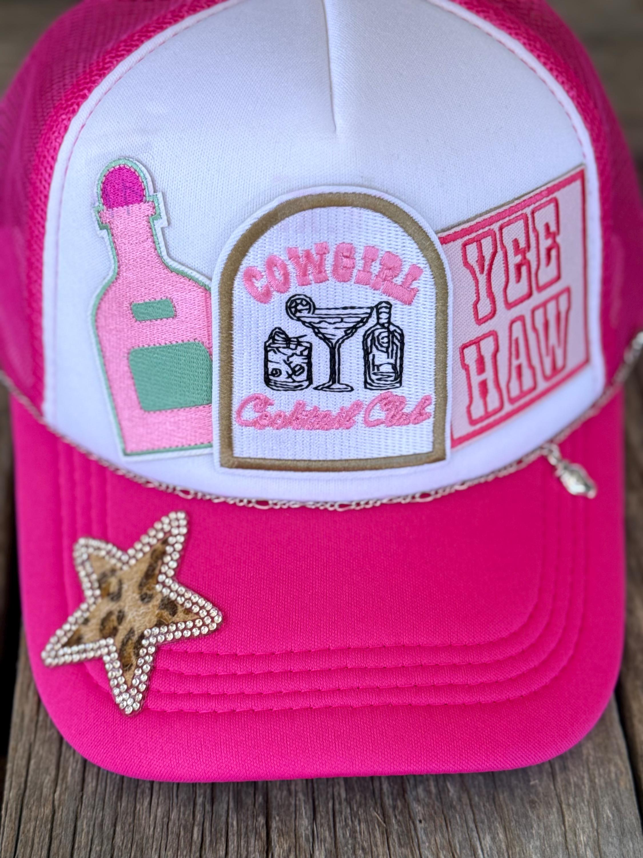 A pink and white trucker hat with embroidered patches, including a 'Cowgirl Cocktail Club' emblem and a 'Yee Haw' patch, plus a rhinestone-embellished star on the brim, set against a rustic wooden background.