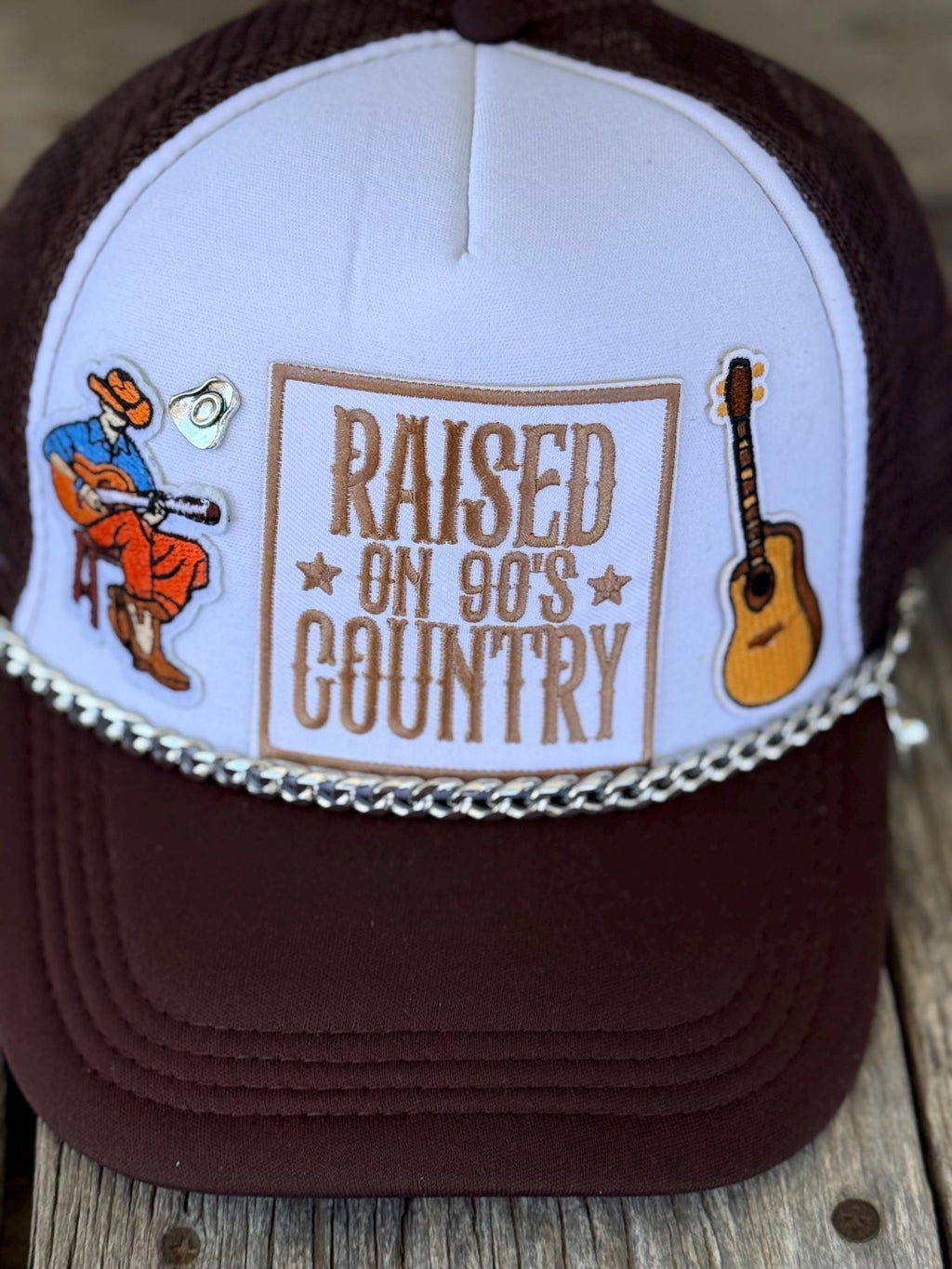 A maroon and white trucker hat with a western-themed design. The front panel is white with a brown embroidered patch reading 'RAISED ON 90'S COUNTRY' in brown, flanked by embroidered illustrations of a cowboy playing a guitar on the left and an acoustic guitar on the right.