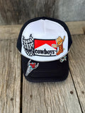 A black and white trucker hat with western-themed patches, including a 'Cowboys' logo, a skeleton cowboy patch, and a checkered flag patch, displayed on a rustic wooden surface.