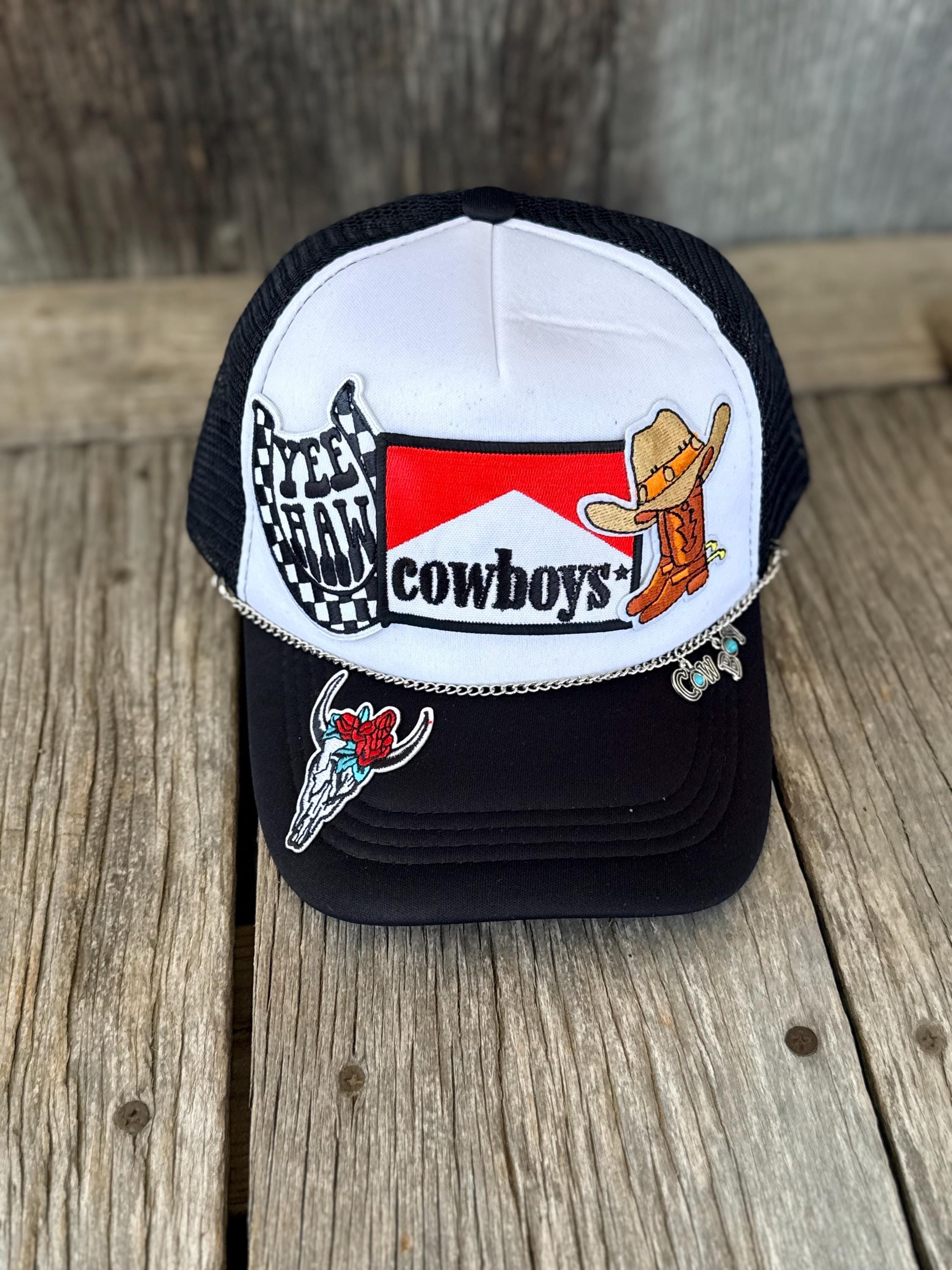 A black and white trucker hat with western-themed patches, including a 'Cowboys' logo, a skeleton cowboy patch, and a checkered flag patch, displayed on a rustic wooden surface.