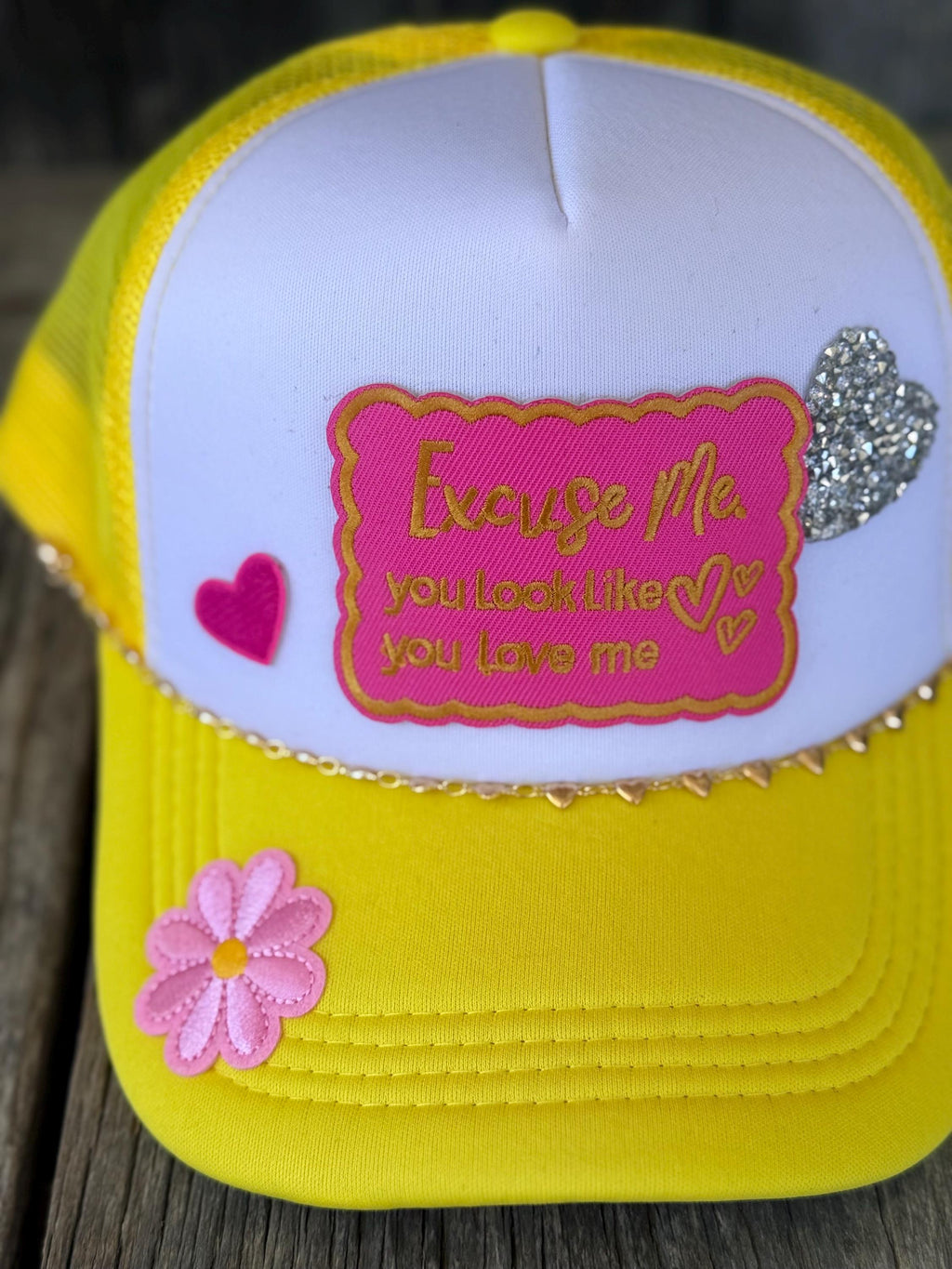A bright yellow and white trucker hat with a pink embroidered patch,' decorated with a pink flower on the brim, a pink heart, and a silver glitter heart.
