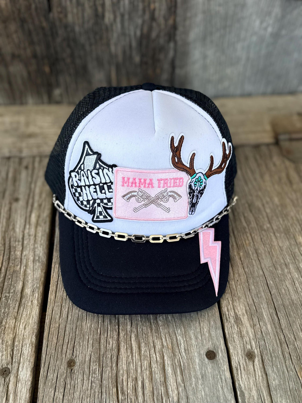 A black and white trucker hat with western-themed patches, including a pink 'MAMA TRIED' patch, a skull with antlers, a lightning bolt, and a chain across the front, displayed on a rustic wooden surface.