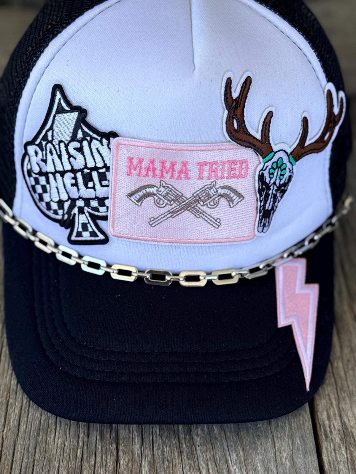 A black and white trucker hat with multiple embroidered patches, including 'RAISING HELL' and 'MAMA TRIED' with crossed guns, a deer skull patch, a pink lightning bolt patch, and a silver chain across the front, shown on a wooden surface.