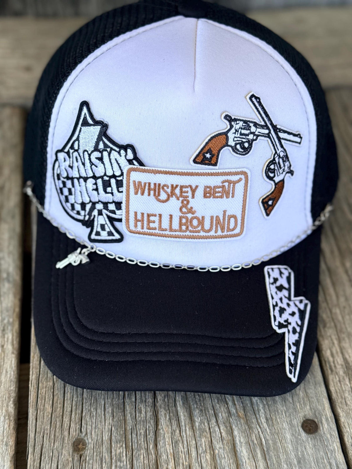 A black and white trucker hat with embroidered patches, including a 'Whiskey' patch and a lightning bolt patch, resting on a rustic wooden surface.