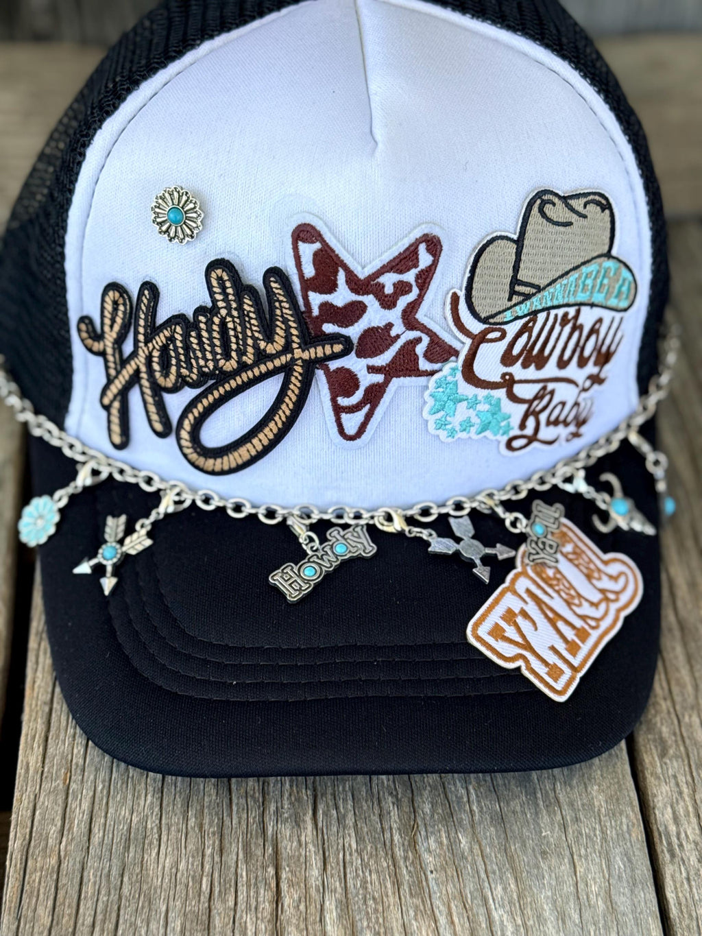 A black and white trucker hat with western embroidery, featuring the words 'Howdy' and a cowboy hat patch, adorned with a silver chain and turquoise conchos resting on a wooden surface.