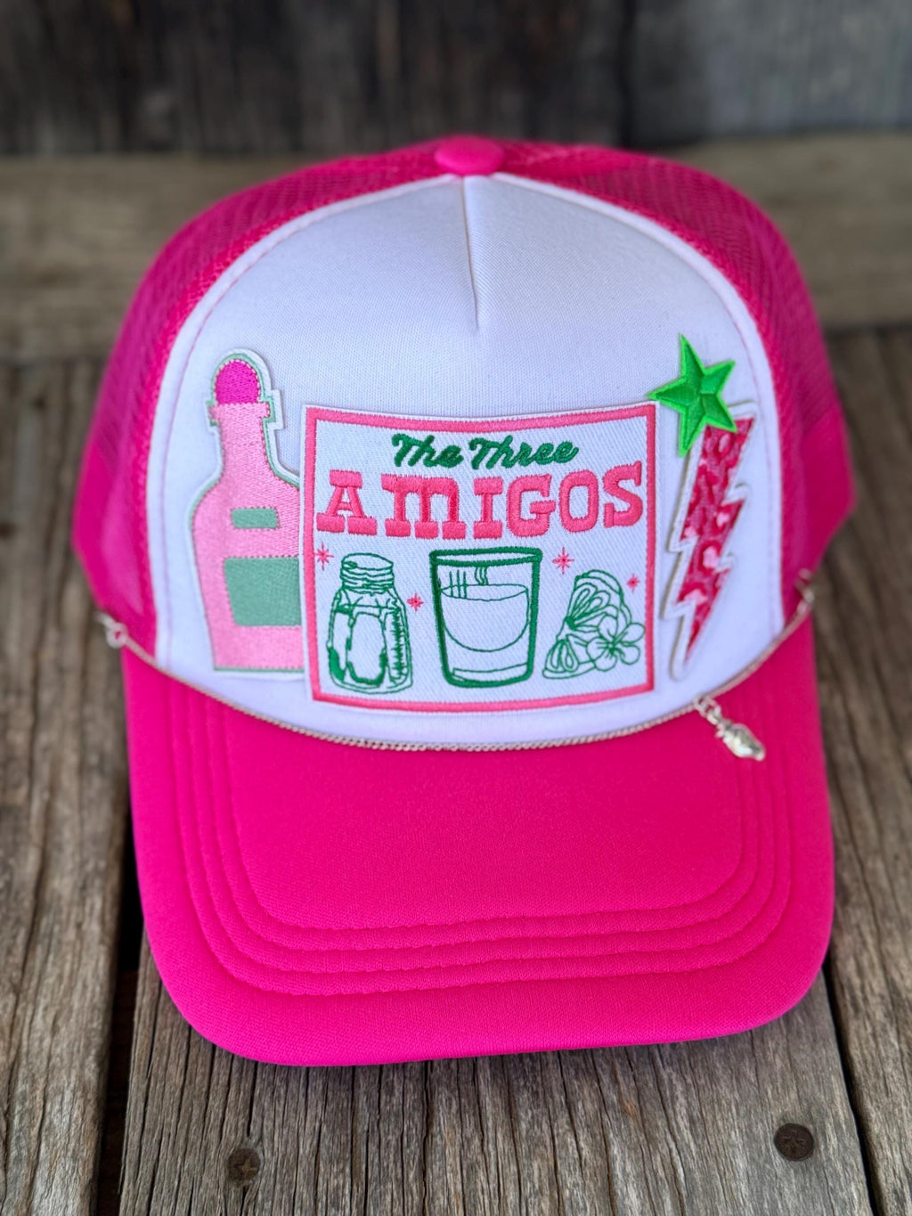 A hot pink and white trucker hat with a custom embroidered front patch reading 'The Three AMIGOS' and featuring illustrations of a tequila bottle, margarita glass, salt shaker, and a red chili pepper, displayed on a rustic wooden surface.