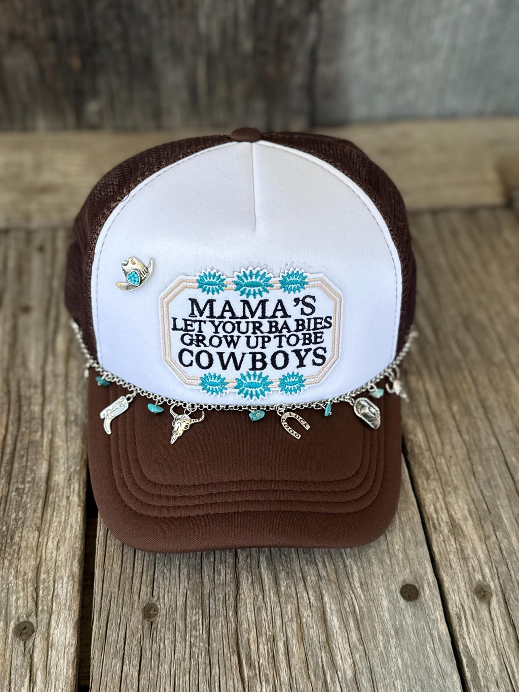 A brown and white trucker hat with a western-themed embroidered design on the front panel with turquoise floral accents. The hat is decorated with a silver chain featuring cowboy and horse charms, turquoise beads, and a small cowboy hat charm, resting on a rustic wooden surface.