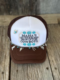 A brown and white trucker hat with a western-themed embroidered design on the front panel with turquoise floral accents. The hat is decorated with a silver chain featuring cowboy and horse charms, turquoise beads, and a small cowboy hat charm, resting on a rustic wooden surface.