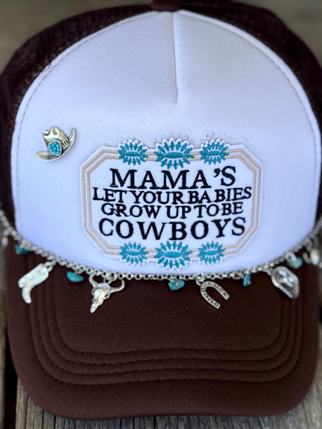A close-up of a brown and white trucker hat with embroidered cowboy patch on the front panel. The hat has turquoise and silver decorative embroidery, dangling cowboy-themed charms, and a small cowboy hat pin on the side.