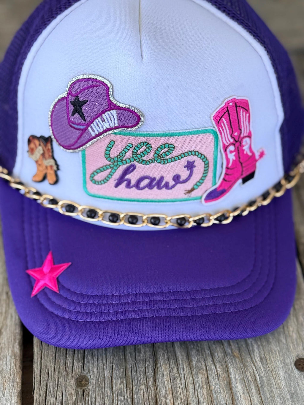 A purple and white trucker hat with western-themed embroidery, including a cowboy hat, boots, and the words 'yee haw', plus a gold chain across the front and a pink star on the brim, shown on a rustic wooden surface.