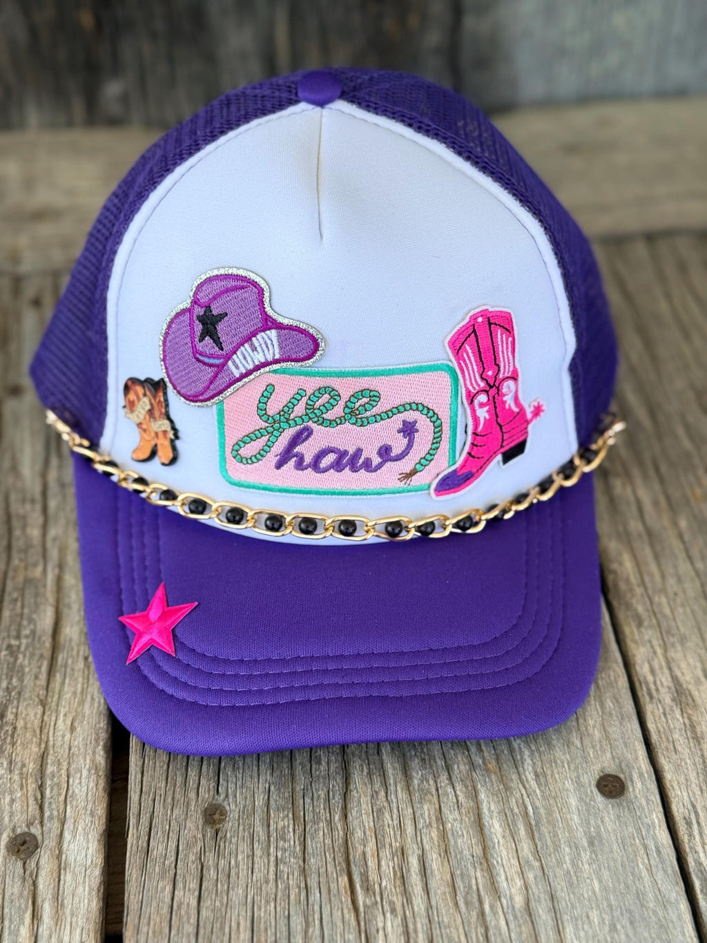 A purple and white trucker hat with western-themed embroidery, including a cowboy hat, boots, and the text 'hee haws', resting on a rustic wooden surface.
